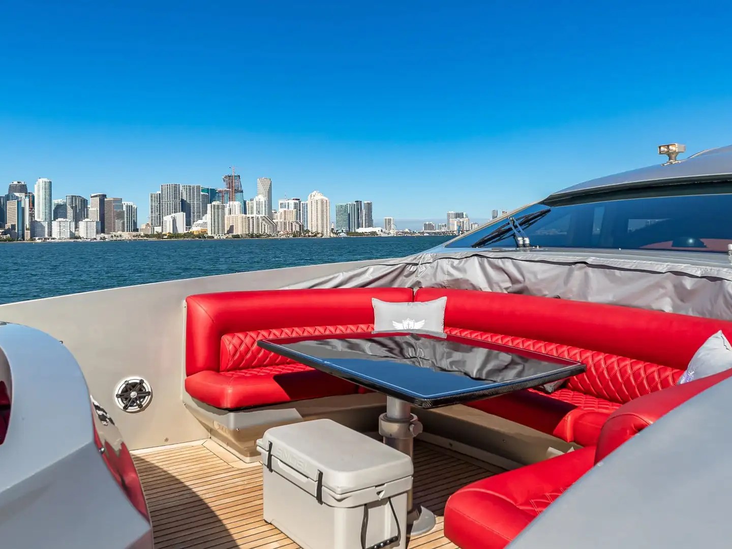 90 Pershing Yacht Charter forward lounge with red wraparound seating, cocktail table, teak decking, and Downtown Miami skyline views across South Florida waters