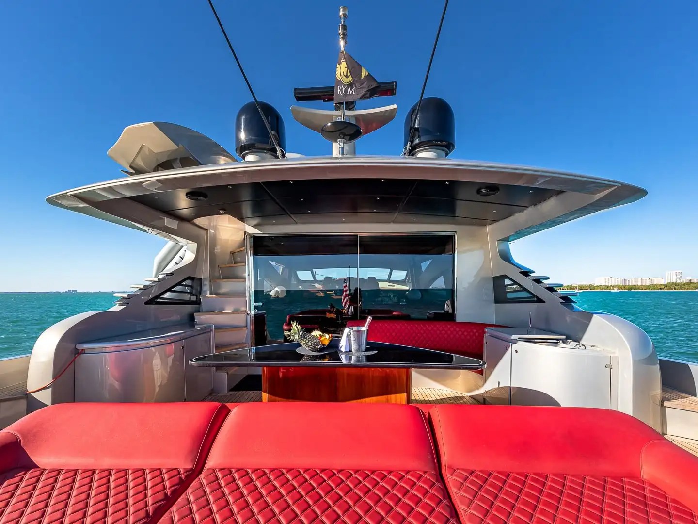 90 Pershing Yacht Charter aft deck viewed from sun lounge featuring red diamond-stitched seating, outdoor dining table, and Miami coastal waters near Key Biscayne
