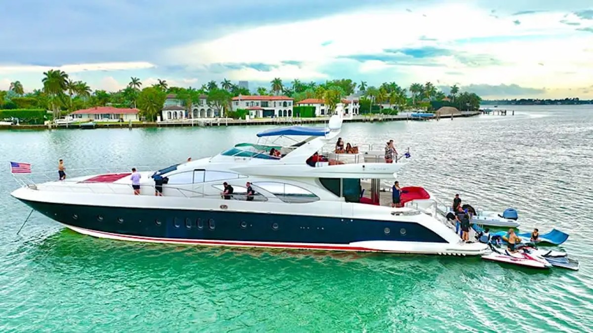 Luxury yacht on turquoise water, people aboard, houses in background.