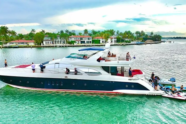 Luxury yacht on turquoise water, people aboard, houses in background.
