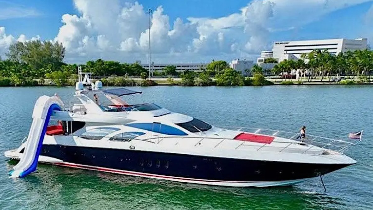 Luxury yacht with a slide docked on a sunny day, trees and buildings in the background.