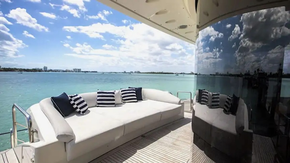 Modern yacht deck with white sofa, striped pillows, and ocean view under a partly cloudy sky.