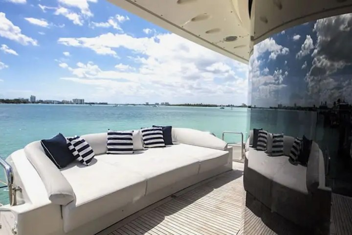 Modern yacht deck with white sofa, striped pillows, and ocean view under a partly cloudy sky.