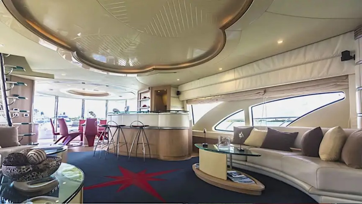 Luxurious yacht interior with sofas, glass table, bar stools, and large windows on a sunny day.
