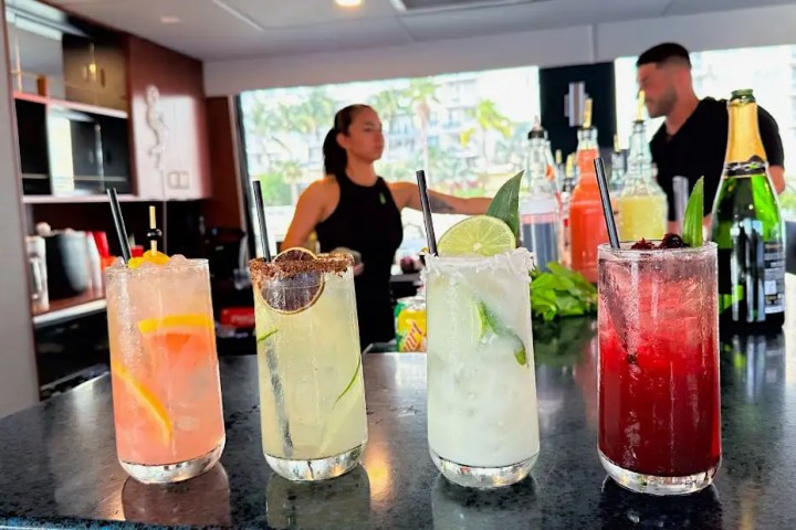 Four colorful cocktails lined up on a bar with two people in the background.