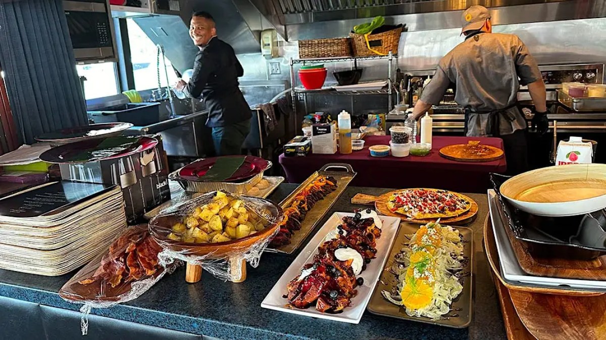 Chefs preparing food in a kitchen with dishes of fruit, bacon, steak, and salad displayed on a counter.