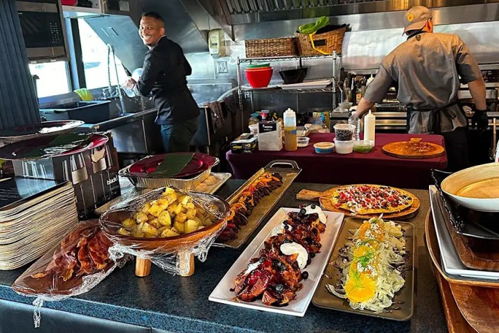 Chefs preparing food in a kitchen with dishes of fruit, bacon, steak, and salad displayed on a counter.