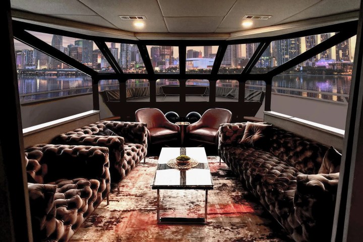 Luxurious yacht interior with tufted sofas, chairs, table, and city skyline view through large windows.