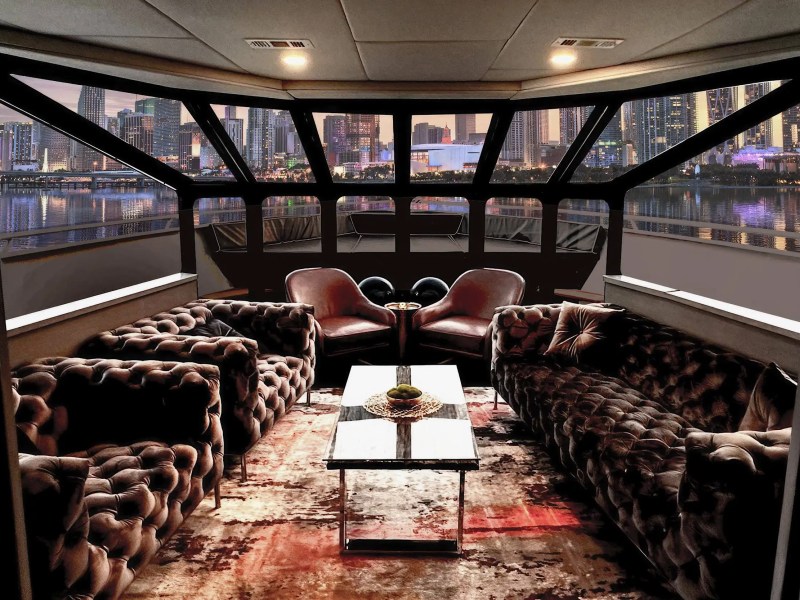 Luxurious yacht interior with tufted sofas, chairs, table, and city skyline view through large windows.