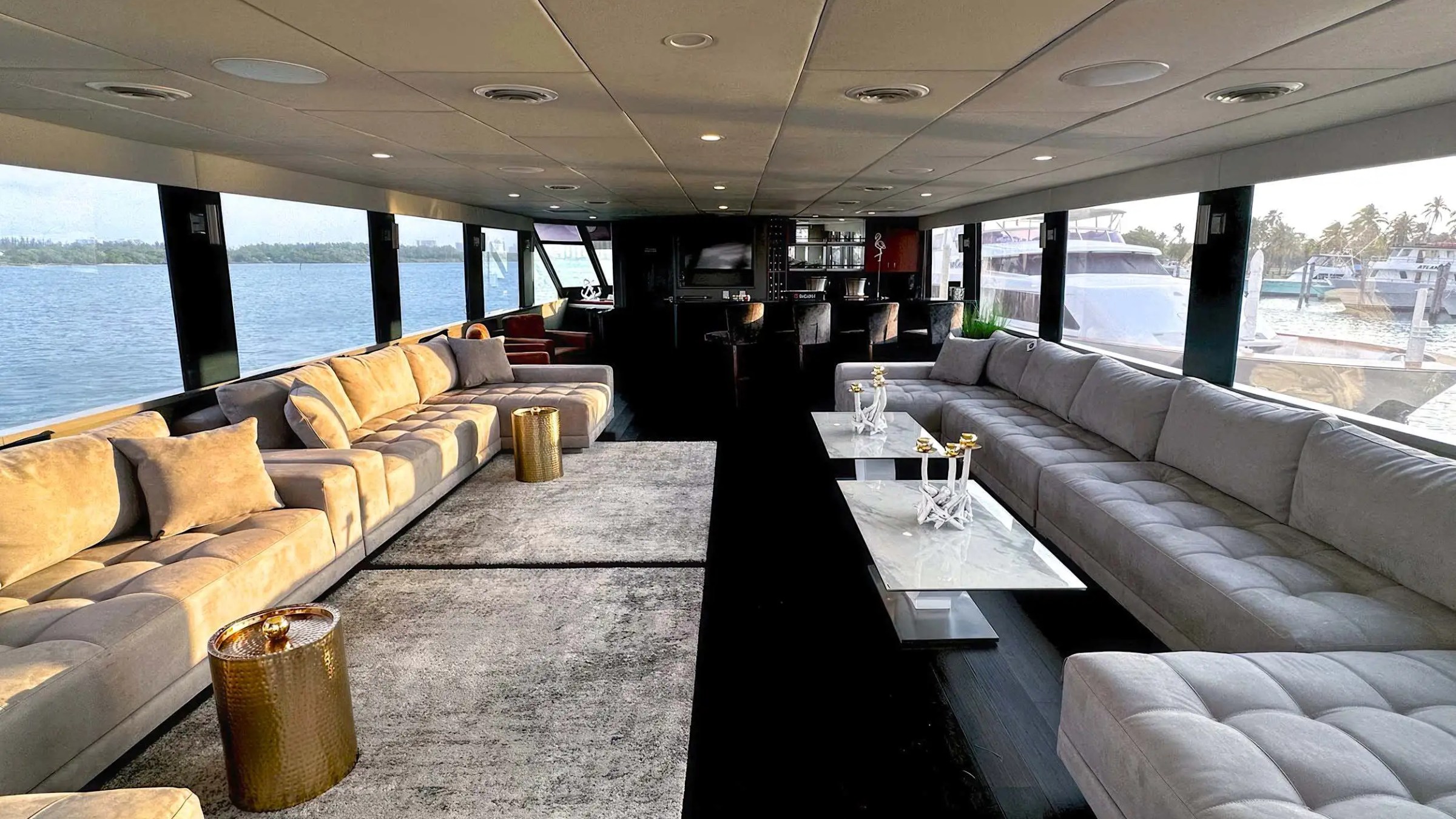 Luxurious yacht interior with grey sofas, gold tables, and large windows overlooking water.