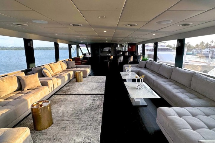 Luxurious yacht interior with grey sofas, gold tables, and large windows overlooking water.