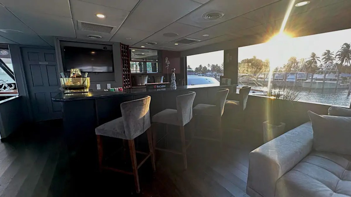 Bar interior with seating, TV, and sunlight through large windows overlooking water and palm trees.