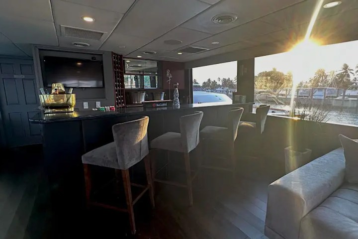 Bar interior with seating, TV, and sunlight through large windows overlooking water and palm trees.