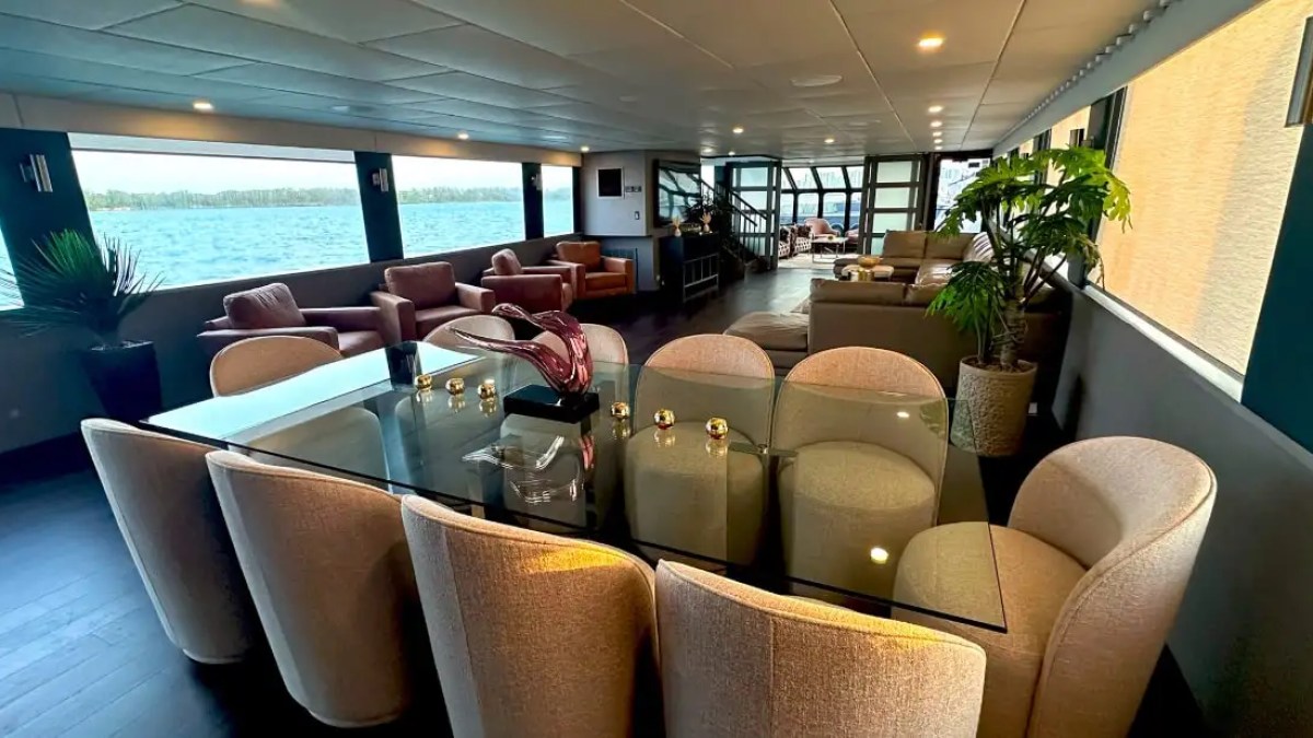 Spacious ship interior with glass dining table, chairs, sofas, and large windows overlooking water.