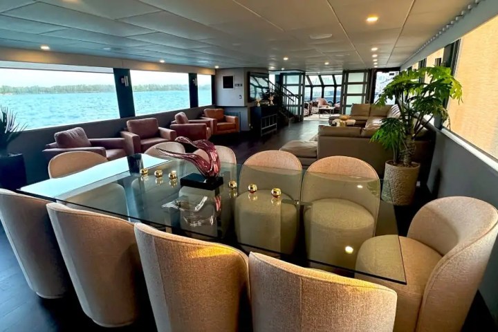 Spacious ship interior with glass dining table, chairs, sofas, and large windows overlooking water.