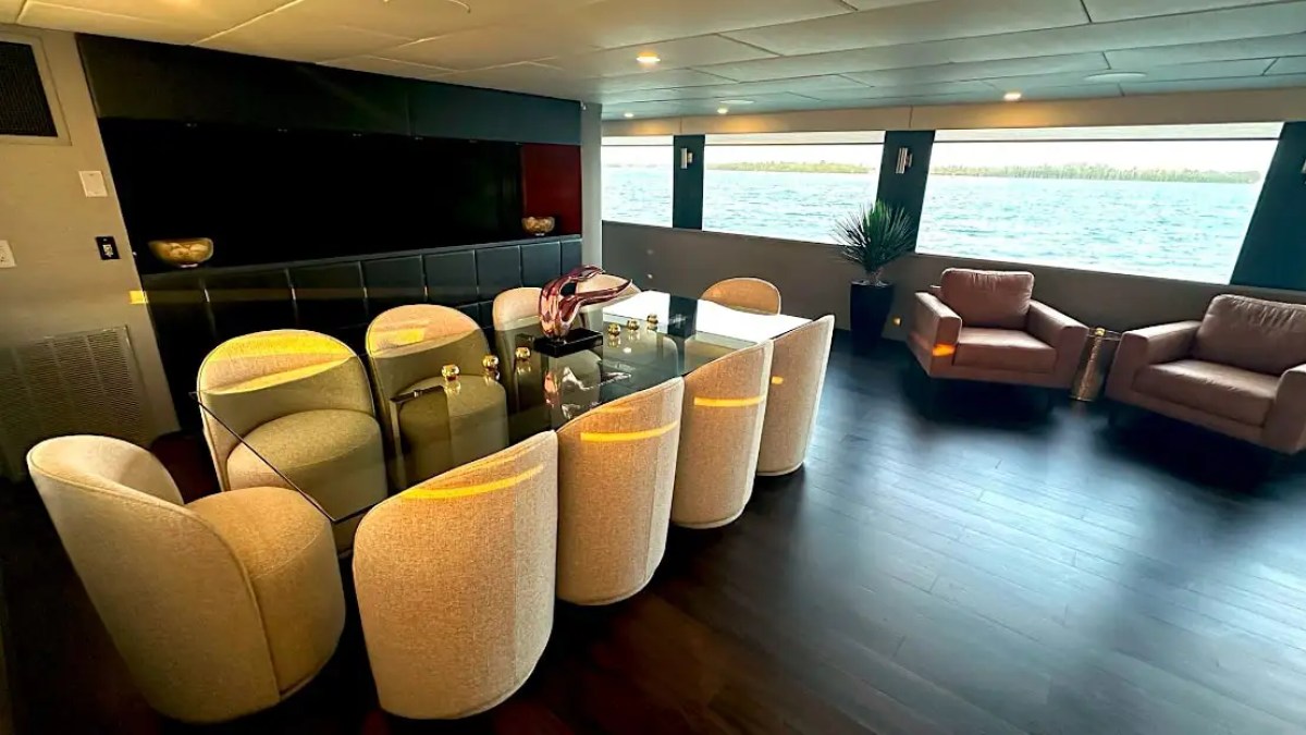 Luxurious yacht interior with dining table, chairs, armchairs, and sea view through large windows.