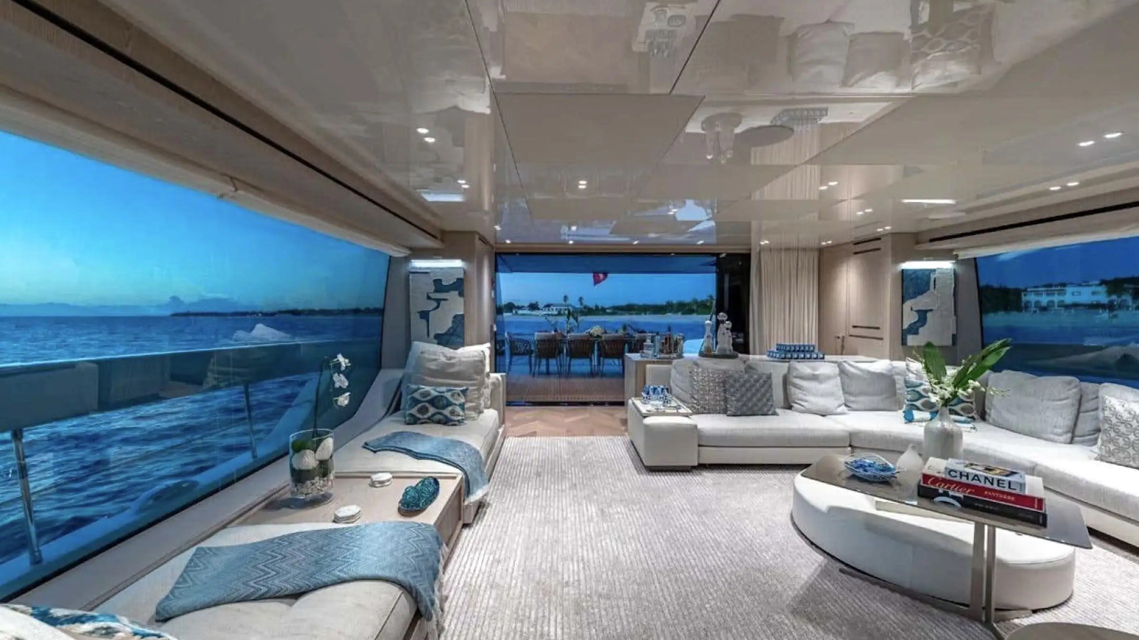 Luxurious yacht interior with white sofas, blue accents, large windows, and ocean view.