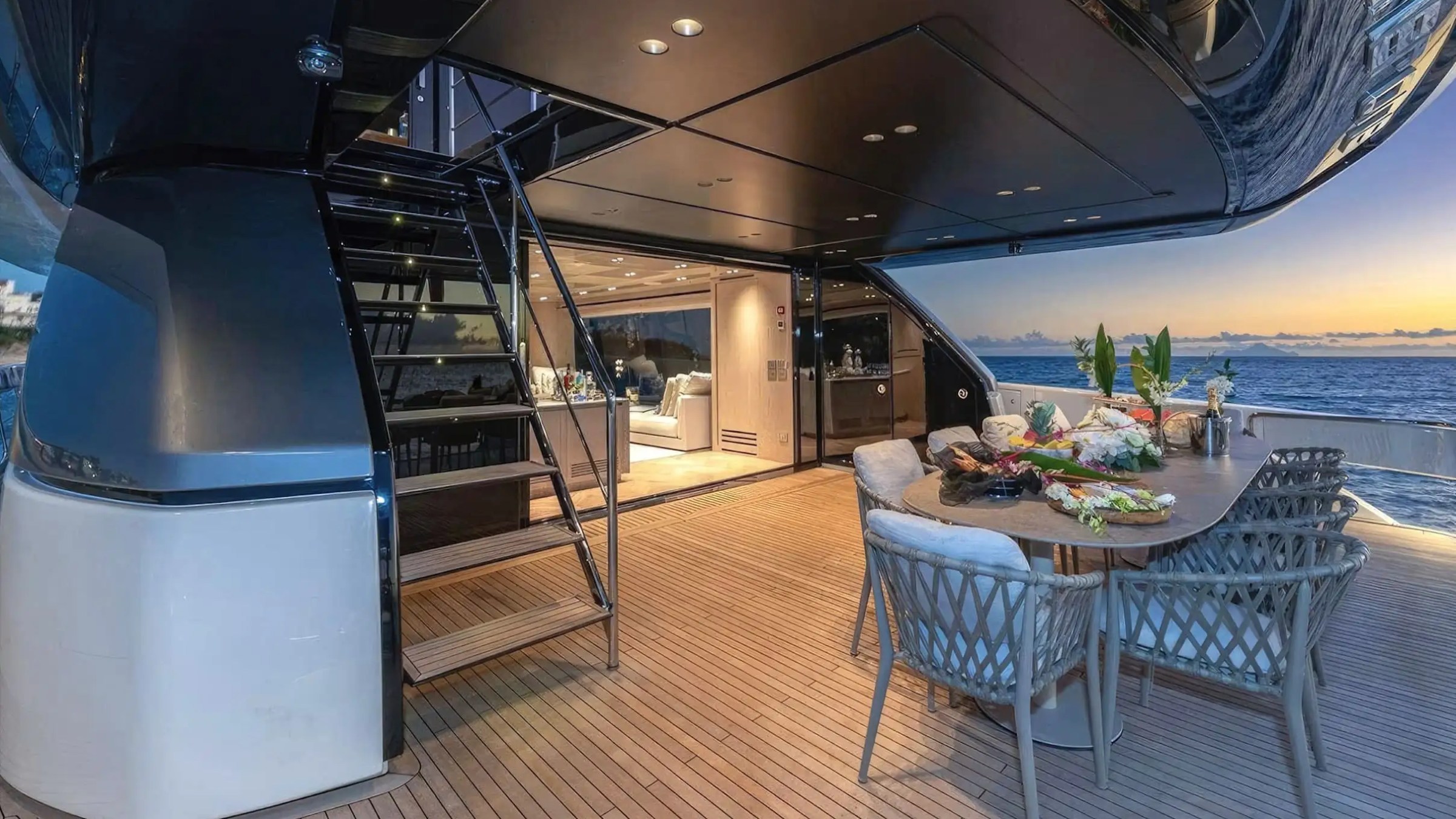 Luxurious yacht deck with dining table, staircase, and ocean view at sunset.