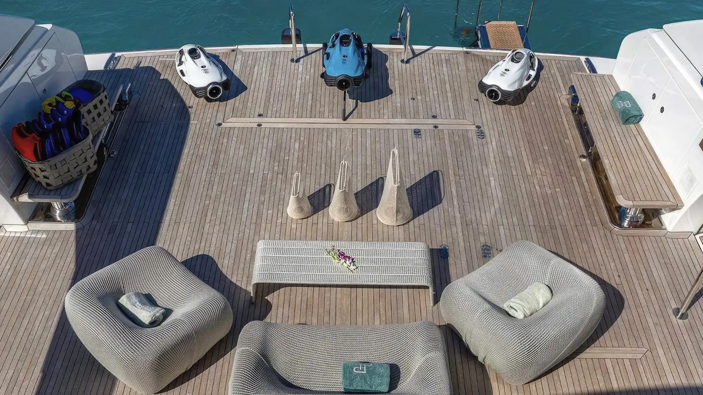 Yacht deck with seating, table, diving gear, and three underwater scooters; view of water in the background.