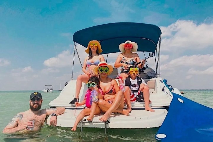 Five people in swimwear on a boat, wearing hats and novelty sunglasses, with a bright blue sky as the backdrop.