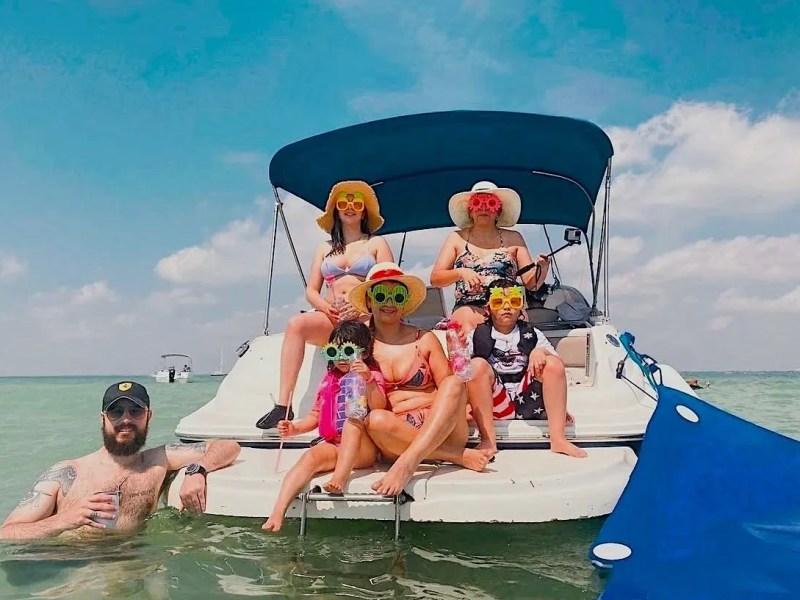 Five people in swimwear on a boat, wearing hats and novelty sunglasses, with a bright blue sky as the backdrop.