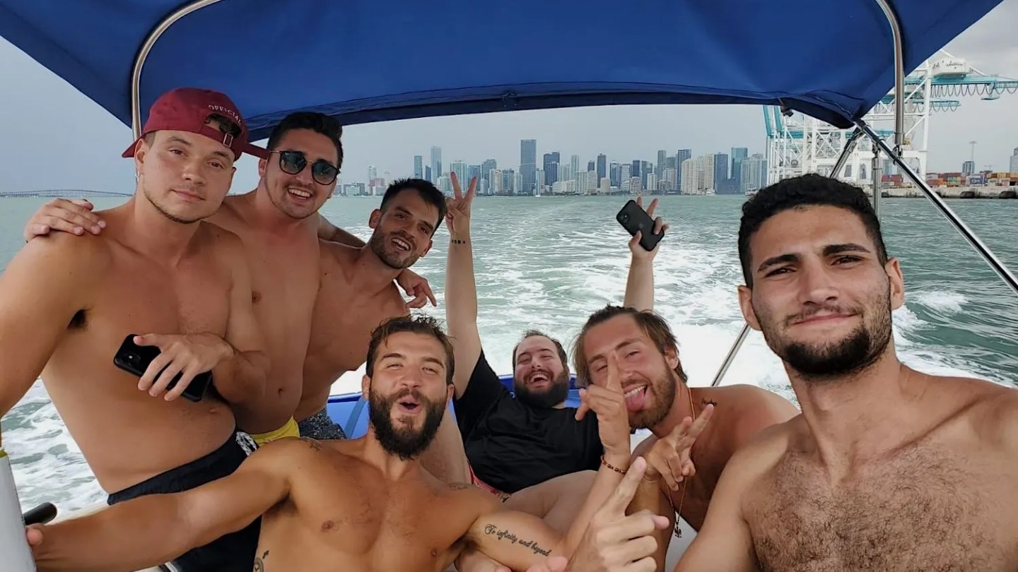 Seven shirtless men on a boat smiling, with a city skyline in the background.