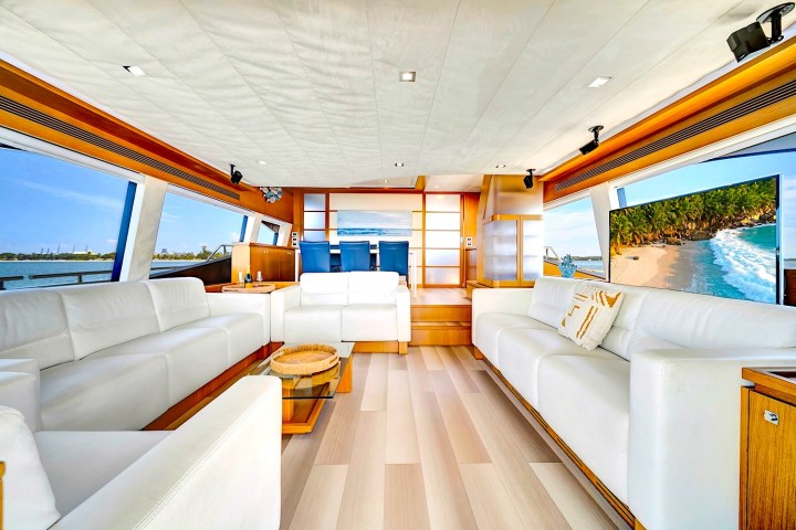 Luxurious yacht interior with white sofas, wooden accents, large windows, and a wall-mounted TV showing a beach scene.