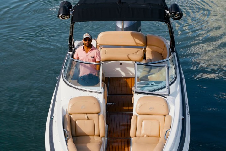 Man steering a boat with tan seats and dark canopy on calm water.