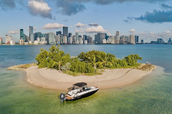 Small boat near sandy island with trees, city skyline and clouds in the background.