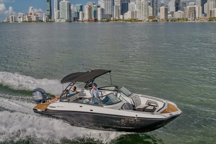 Boat with two people cruising on water, city skyline in background under blue sky.