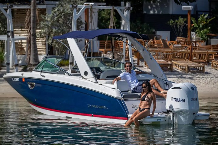 Man and woman relaxing on a motorboat near a sandy beach with lounge chairs.