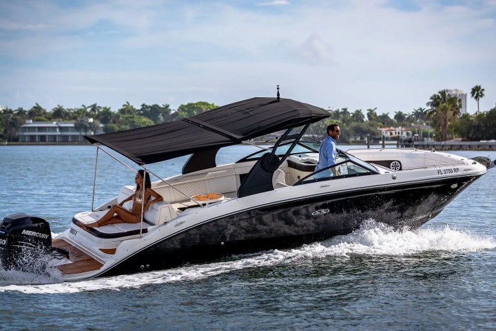 Powerboat with two people cruising on a sunny day with trees and houses in the background.