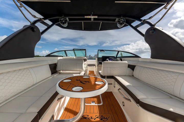 Interior view of a motorboat with white seats and a wooden table, under a canopy on a sunny day.