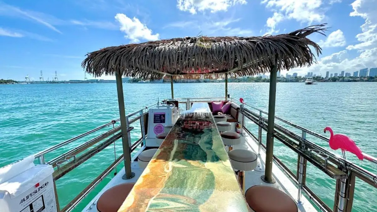 View from a tropical-themed boat with a bar, stools, flamingo decorations, and city skyline in the background.