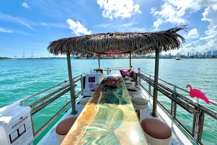 View from a tropical-themed boat with a bar, stools, flamingo decorations, and city skyline in the background.