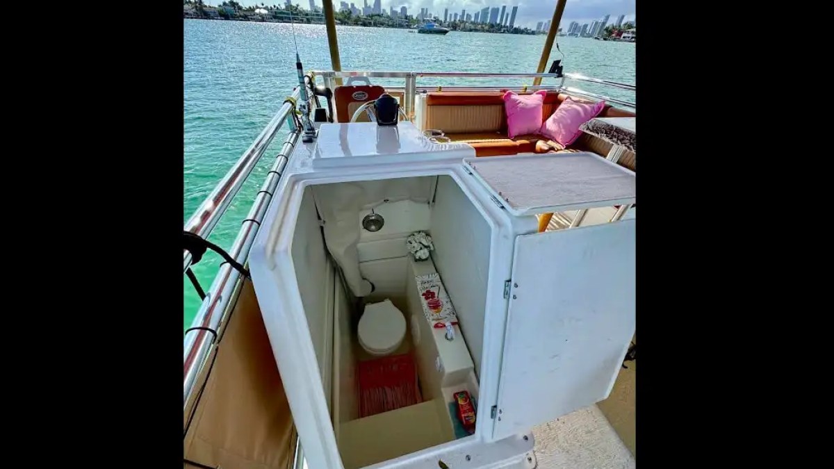 Small boat deck with open hatch revealing a compact toilet, seating area with pink cushions, and city skyline.