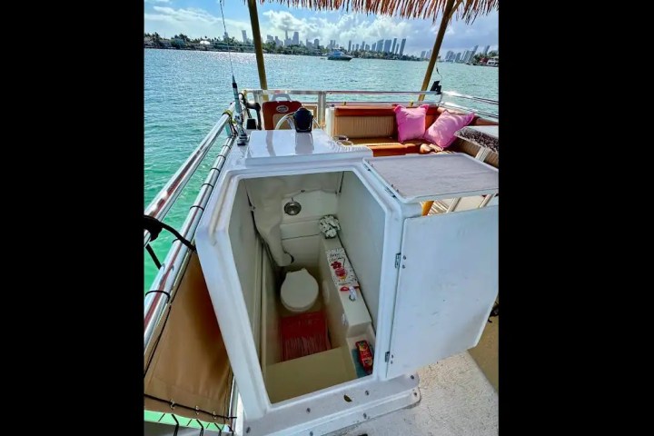 Small boat deck with open hatch revealing a compact toilet, seating area with pink cushions, and city skyline.