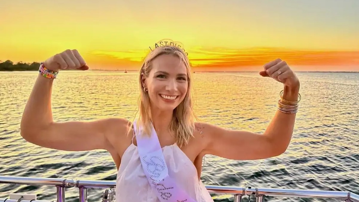 Smiling woman flexing arms on a boat at sunset, wearing a sash and tiara.