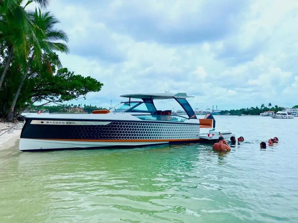 A 32 Saxdor Boat Rental anchored beside a tropical Miami Beach shoreline with guests swimming in shallow clear water, showcasing Scandinavian styling and luxury day boating in South Florida