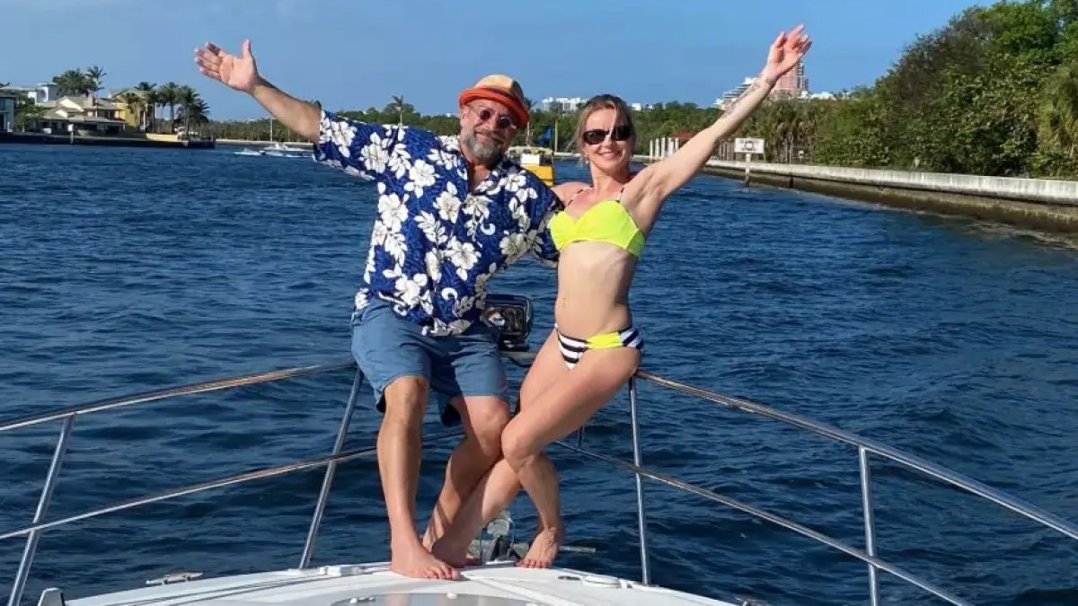 Two people in swimwear pose joyfully on a boat with open arms, surrounded by water and trees.