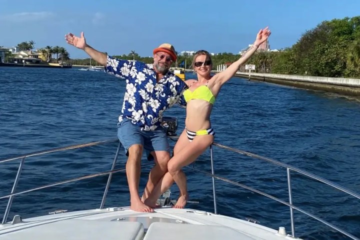Two people in swimwear pose joyfully on a boat with open arms, surrounded by water and trees.
