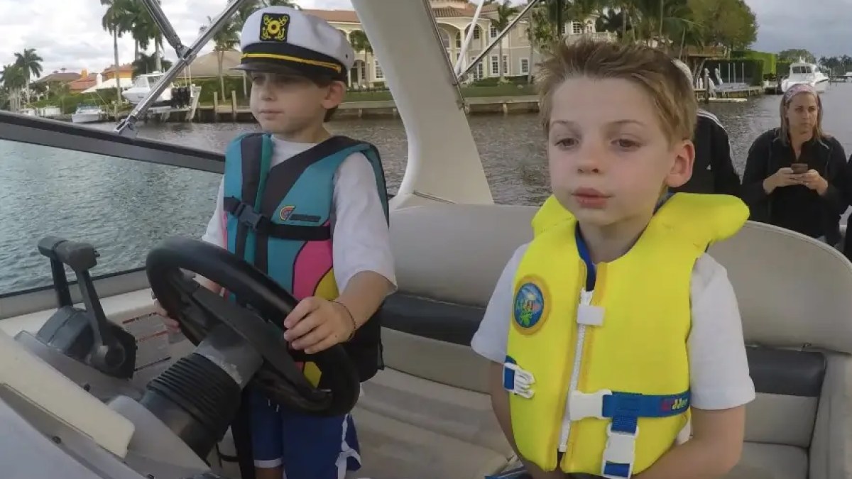 Two boys on a boat, wearing life jackets; one steering, the other observing.