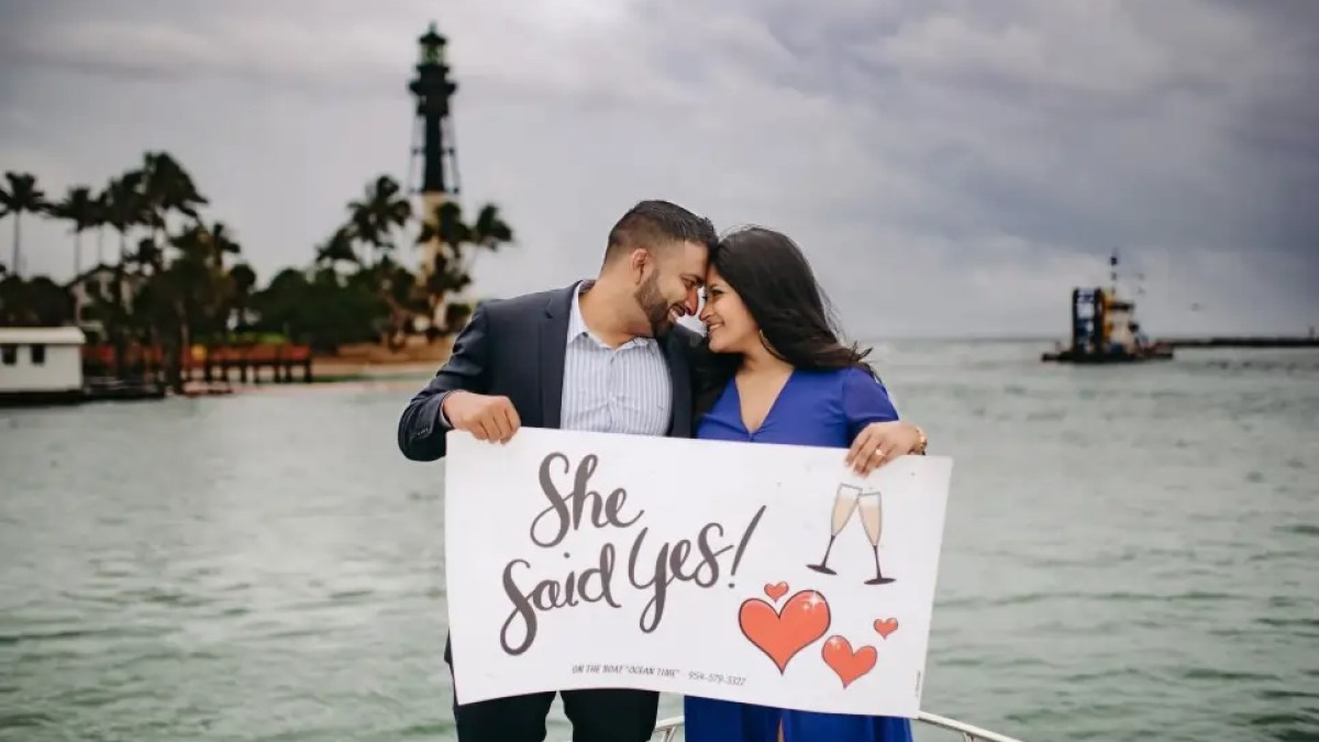 Couple on a boat holding a 'She Said Yes!' sign with a lighthouse in the background.