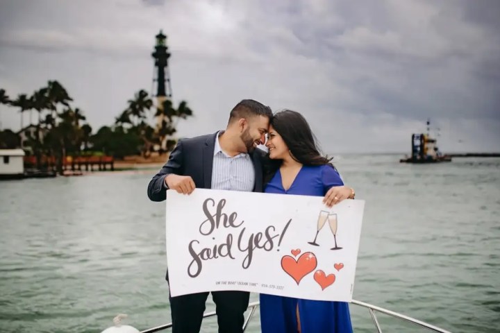 Couple on a boat holding a 'She Said Yes!' sign with a lighthouse in the background.