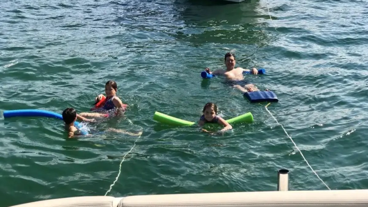 Four people floating with pool noodles and inflatables near a boat on a sunny day.