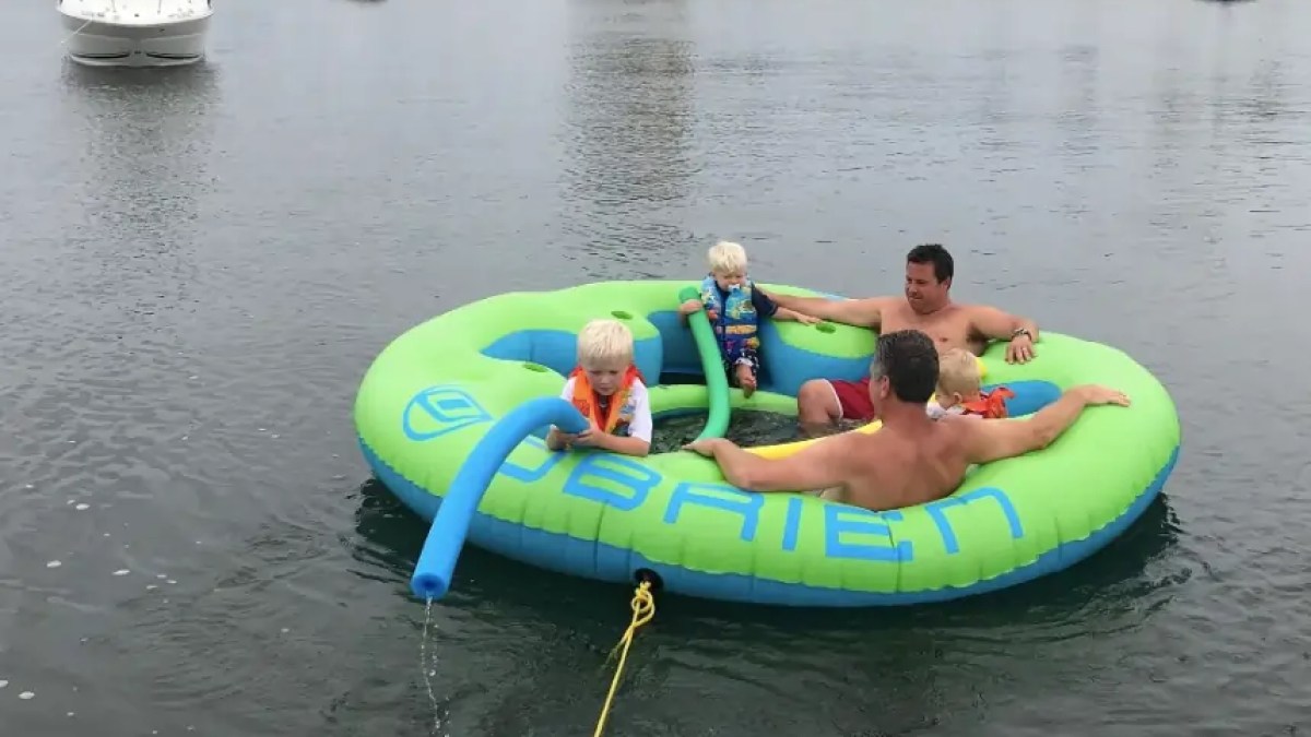 Two adults and two children on a large inflatable tube in a body of water with boats in background.