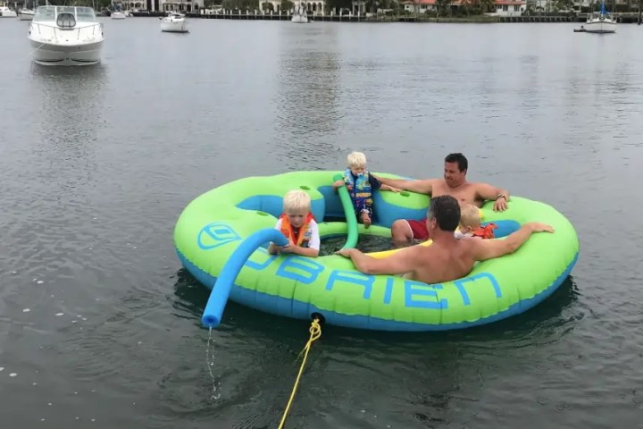 Two adults and two children on a large inflatable tube in a body of water with boats in background.