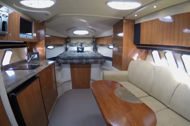 Interior of a yacht with a bed, kitchenette, and sofa in a wood and white color scheme.