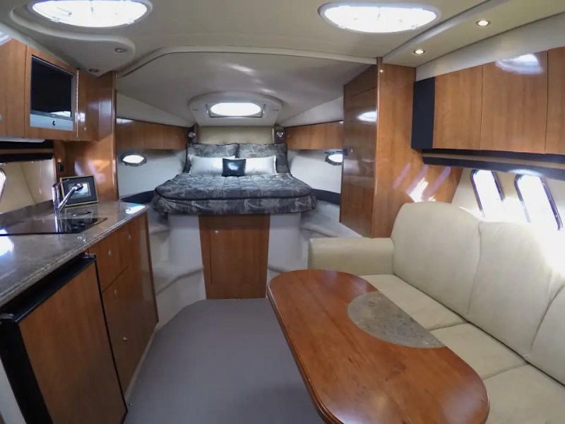 Interior of a yacht with a bed, kitchenette, and sofa in a wood and white color scheme.