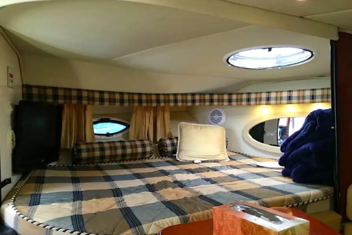 Cozy boat cabin with a plaid bed, pillows, circular windows, and a tissue box on a wooden table.